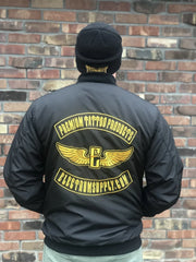 A person wearing a black Electrum Supply bomber jacket with a gold and white logo on the back, standing in front of a brick wall.