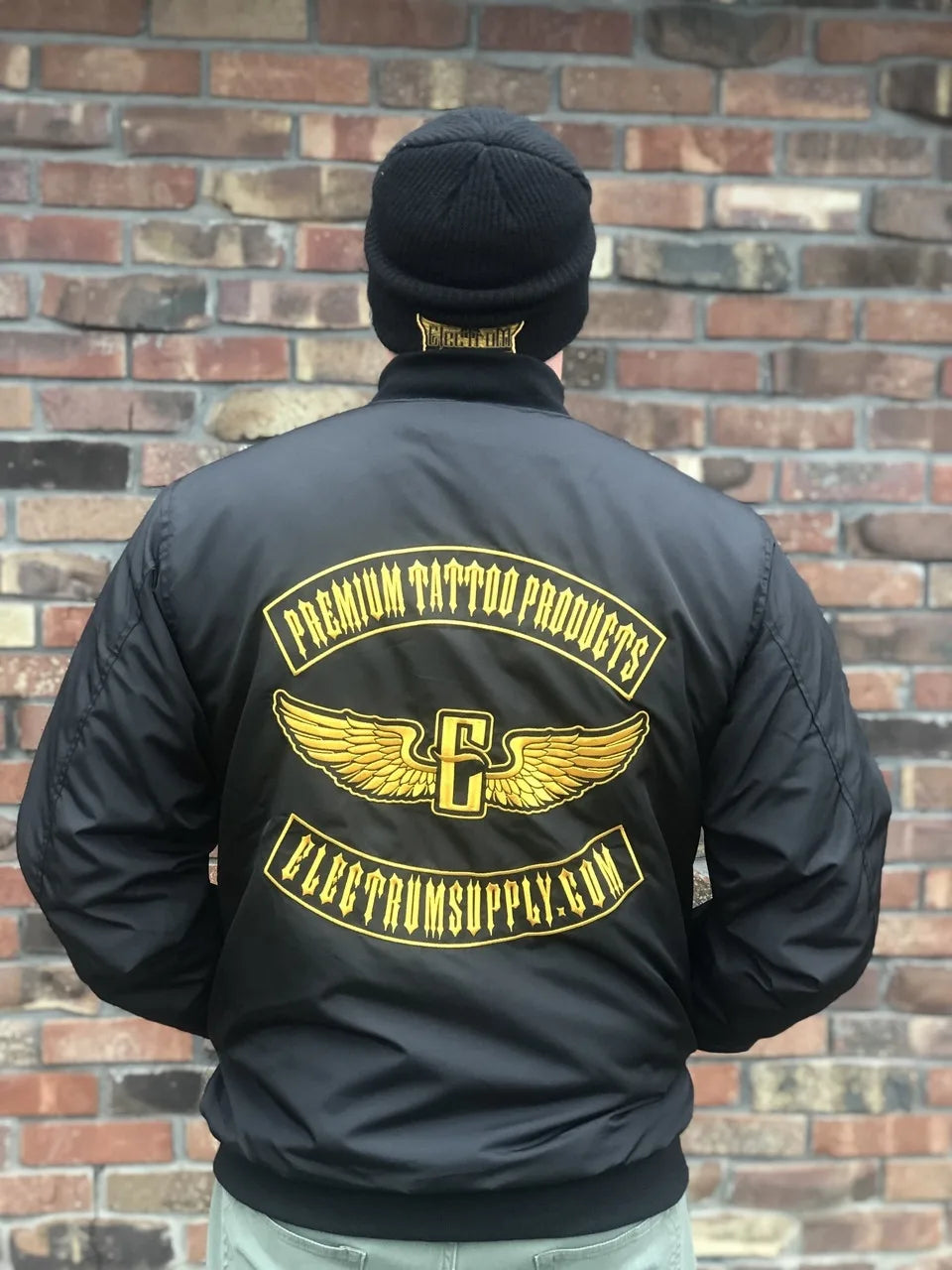 A person wearing a black Electrum Supply bomber jacket with a gold and white logo on the back, standing in front of a brick wall.