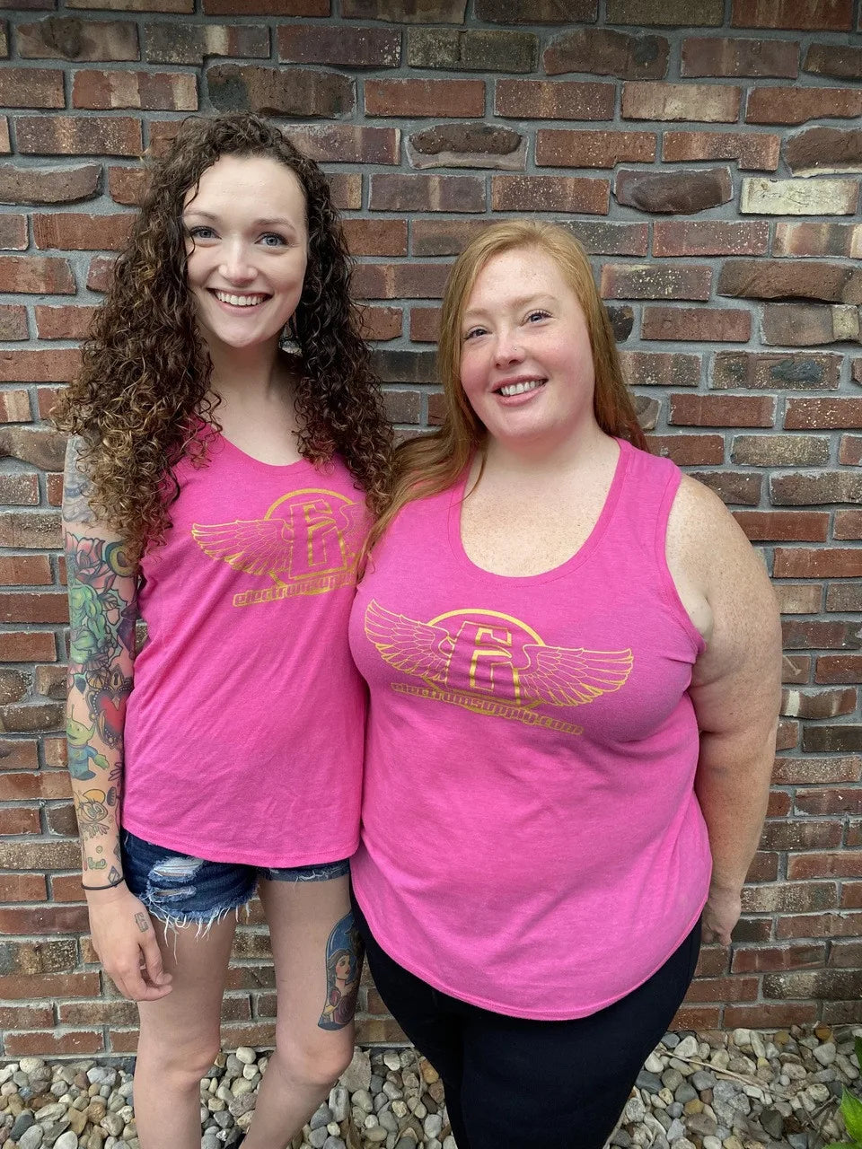 Two individuals wearing pink tank tops with the electrum flying e logo on the front, standing in front of a brick wall.