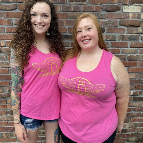 Two individuals wearing pink tank tops with the electrum flying e logo on the front, standing in front of a brick wall.